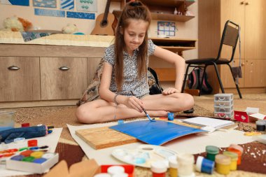 Kız evde resim çiziyor, sanat eseri yapıyor, kağıt, boya ve fırçalardan yaratıcı sanat eserleri yapıyor.