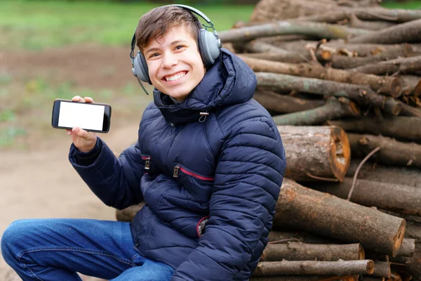 10代の若者はヘッドフォンで音楽を聴き 秋の都市公園でリラックスし 電話画面を見せ 丸太の上に座って — ストック写真