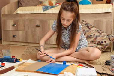 Kız evde resim çiziyor, sanat eseri yapıyor, kağıt, boya ve fırçalardan yaratıcı sanat eserleri yapıyor.