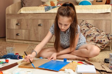 Kız evde resim çiziyor, sanat eseri yapıyor, kağıt, boya ve fırçalardan yaratıcı sanat eserleri yapıyor.