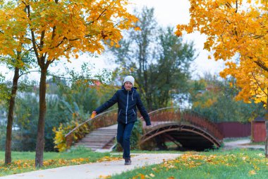 Bir kız yol boyunca yürür ve sonbaharın tadını çıkarır. Sarı yapraklı akçaağaçlar ve nehir üzerinde bir köprü.