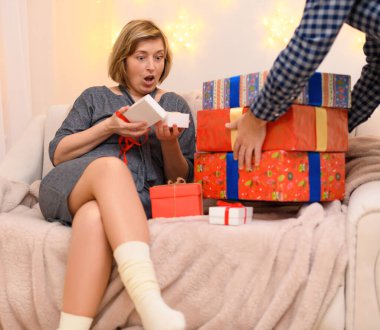 Çok şaşırmış bir kadının portresi, kanepeye oturur ve hediyeleri açar, duvardaki tatil ışıklarını.