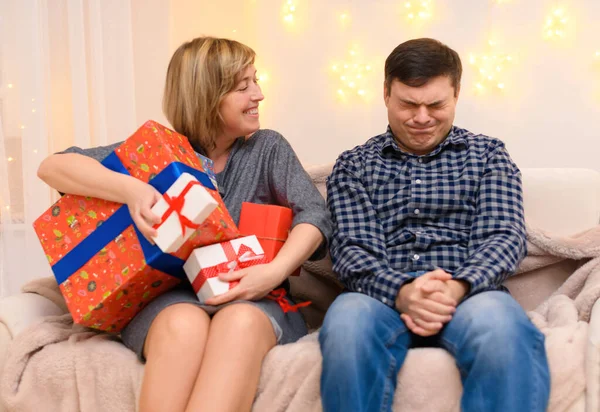 portrait of adult romantic couple, sitting with gifts on a couch at home, a woman takes away all the gifts from a man, the photo is like a joke, holiday lights on a wall