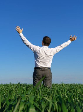 İş adamı bir tarlada poz veriyor, uzaklığa bakıyor ve dinleniyor, arka plan olarak yeşil çimenler ve mavi gökyüzü.