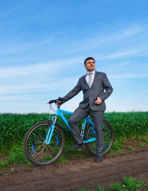 Bisikletli bir işadamı buğday tarlasında poz veriyor, yeşil çimenler ve mavi gökyüzü arka planda