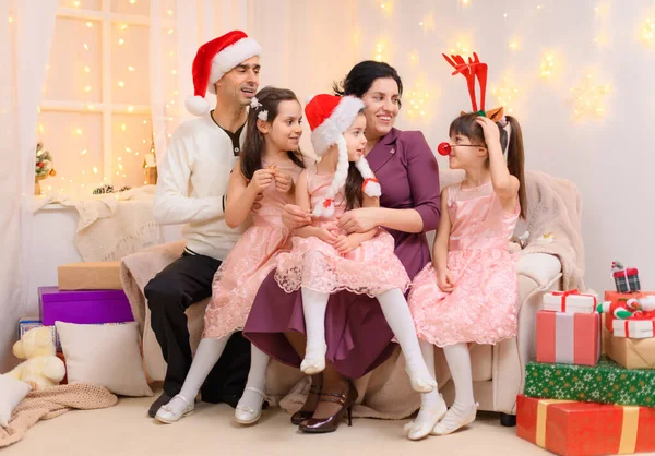 父母和孩子的全家福 头戴圣诞礼帽 坐在家里的沙发上 上面点缀着圣诞彩灯和新年礼物 — 图库照片