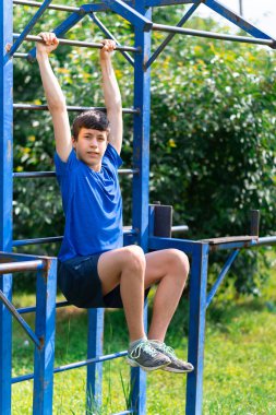 Delikanlı dışarıda spor yapıyor, bahçede spor yapıyor, yatay barda asılı duruyor, bacaklarını kaldırıyor ve karın kaslarını sallıyor, sağlıklı bir yaşam tarzı.