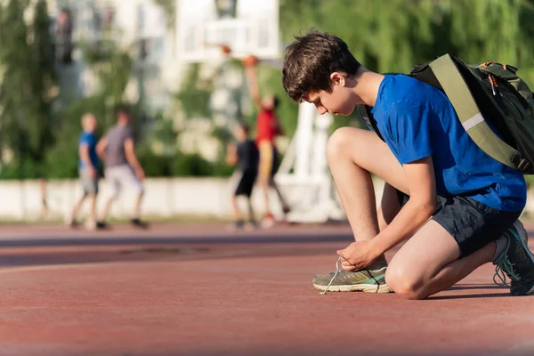 a guy ties his shoelaces on the edge of the field, next to the team playing basketball