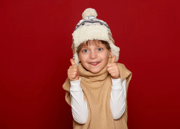 Conceito de inverno - menina em chapéu e suéter mostrar melhor gesto no fundo vermelho — Fotografia de Stock