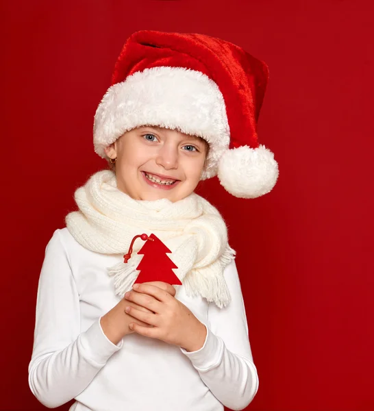 Winter, child,  christmas concept - happy girl in santa hat show fir tree wool toy on red — ストック写真