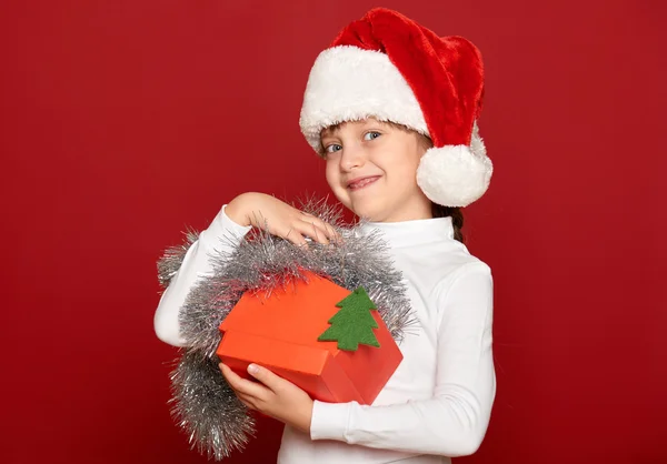 Inverno, criança, conceito de natal - menina feliz em chapéu de santa com caixa de presente mostrar brinquedo de abeto no vermelho — Fotografia de Stock