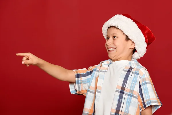 Winterurlaub Weihnachtskonzept - Junge mit Weihnachtsmütze zeigt mit dem Finger auf roten Hintergrund — Stockfoto