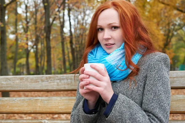 杯茶坐在城市公园的长凳上的红发女孩的秋天 — 图库照片