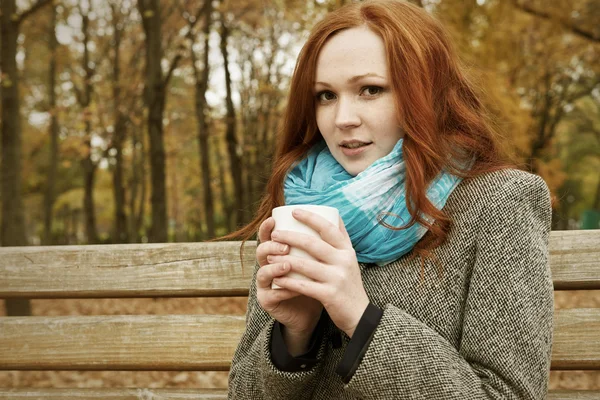 Menina ruiva com xícara de chá sentar em um banco no parque da cidade, temporada de outono — Fotografia de Stock