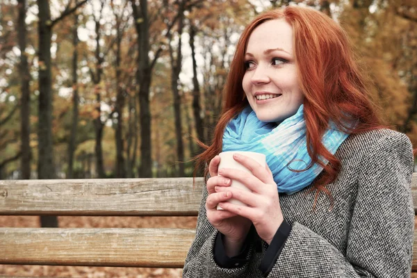 Happy girl with cup of tea sit on a bench in city park, fall season — Stock Photo, Image