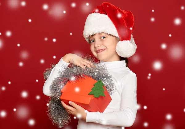 Menina feliz no chapéu com caixa de presente mostrar brinquedo de lã de abeto no fundo vermelho — Fotografia de Stock