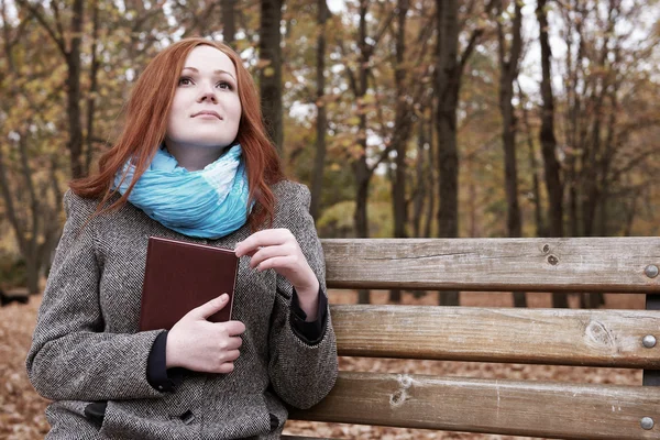 Menina ruiva com livro sentar em um banco no parque da cidade, temporada de outono — Fotografia de Stock