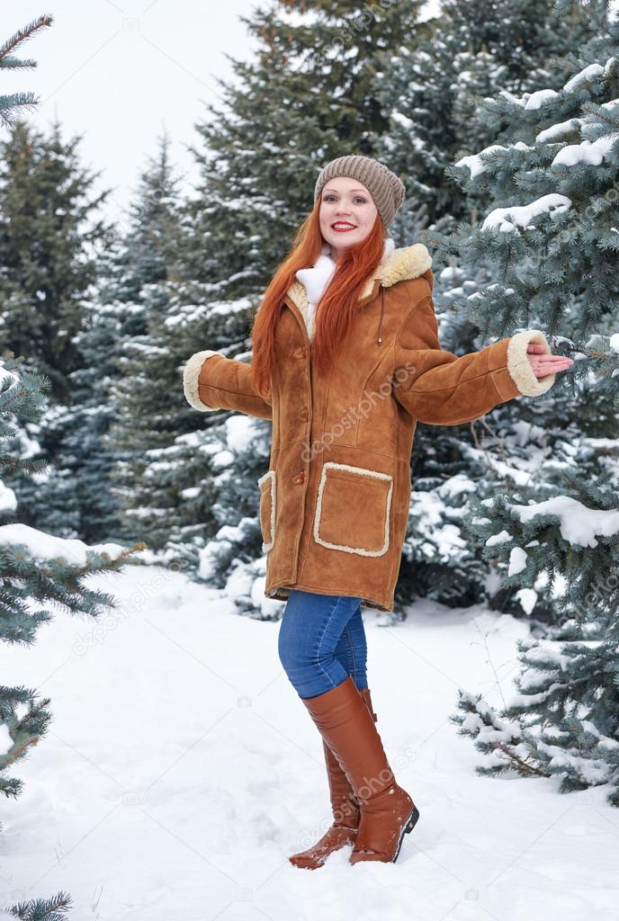 Girl in winter park at day. Fir trees with snow. Redhead woman full length.
