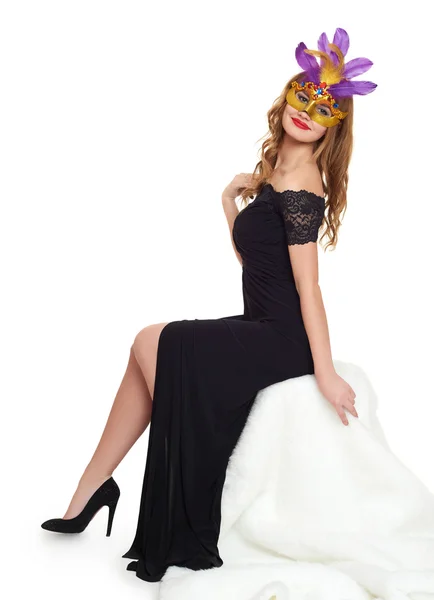 Young woman in black evening gown and carnival mask. Sit on white fur. Holiday and party concept. — Stock Photo, Image