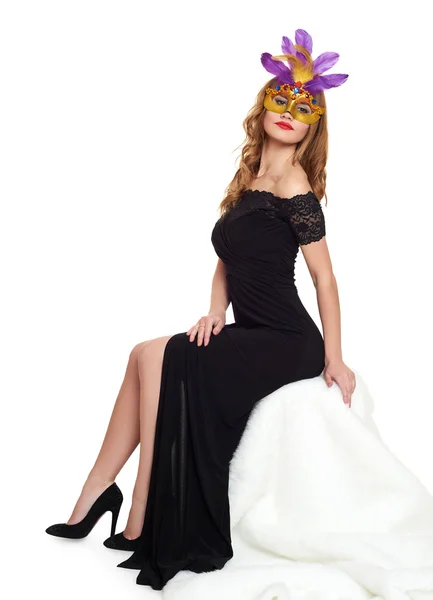 Young woman in black evening gown and carnival mask. Sit on white fur. Holiday and party concept. — Stock Photo, Image