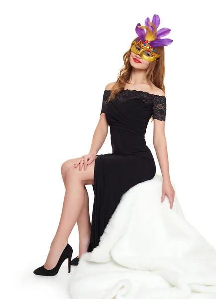 Young woman in black evening gown and carnival mask. Sit on white fur. Holiday and party concept. — Stock Photo, Image
