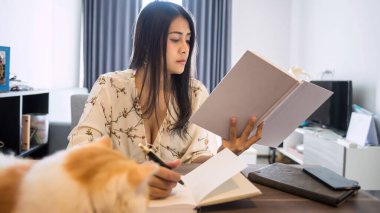 Ciddi Asyalı iş kadını kitap okuyor ve oturma odasındaki iş projesi için kağıda yazıyor. Covid19 salgını sırasında evde kalıp kediyle çalışmak.