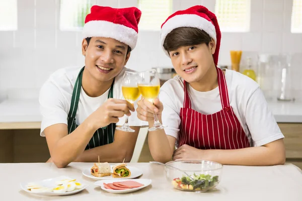 LGBT Asian gay couple with santa hat and apron hold wine and eat salad and sandwich at lunch to celebrate Christmas and 2021 new year after cooking food. Happy together. Same sex family.