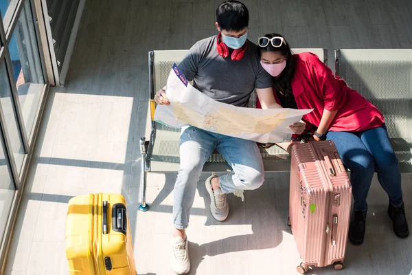 Top view of Asian young couple in face mask and luggage checking transportation subway train map. They wait for airplane departure in airport terminal. Travel new normal to prevent covid-19.