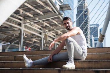 Spor kıyafetleri içinde yakışıklı İtalyan sakallı adam şehir merkezinde egzersiz yaptıktan sonra merdivenlerde dinleniyor ve bacağını esnetiyor. Vücut geliştirme ve sağlıklı yaşam tarzı konsepti. Kasabada spor yapan biri..