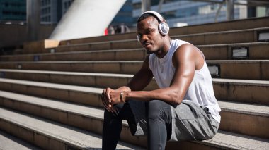 Afrikalı Amerikalı adam spor yaptıktan sonra internetten akıllı telefondan müzik dinliyor. Merdivenlerde rahatça oturuyor. Vücut geliştirme ve egzersiz konsepti. Siyah hayatlar önemlidir.