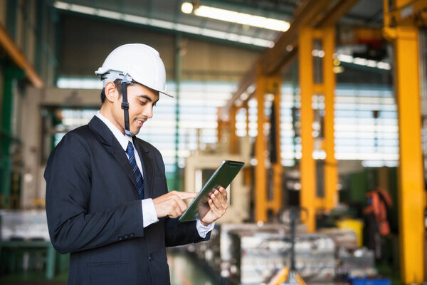 portrait of happy Asian factory manager with suit and helmet holding digital tablet to check stock in warehouse by corporate application. Manufacturing and inventory business concept.