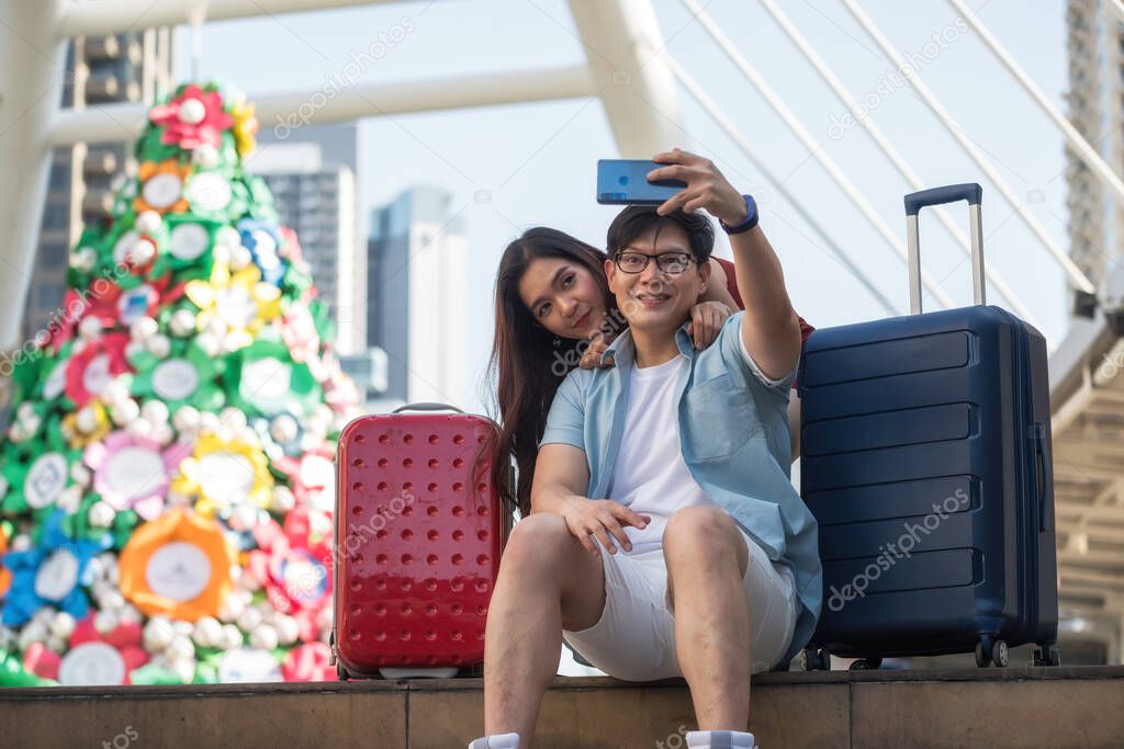 Asian tourist couple selfie photo from smartphone on staircase with two travel luggage in modern urban city at summer. Happy anniversay vacation for sweet man and woman in valentine's day.