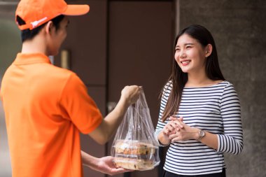 Turuncu üniformalı teslimatçı apartman dairesinin önünde ya da apartmanın önünde gülümseyen kadın müşteriye ekmek fırını veriyor. Covid-19 salgını sırasında çevrimiçi yemek sipariş etmek için yeni normal.