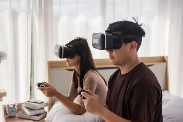 Asian young couple playing game with VR glasses and wireless joystick on bed. They spend time together and relax on weekend. Quarantine, social distancing, stay home to prevent covid-19.