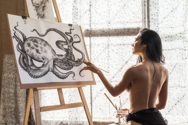 窓から自然光が差し込む紙の板に水彩でタコの絵を描く黒目のアジアのトップレスな若い女性 セクシー女の子でアートギャラリー ストック画像