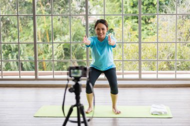 Gülümseyen Asyalı yoga eğitmeni kollarını ve ellerini paspasa uzatıp kameraya bakıyor. Evde video bloğu ve yeşil ağaç çevresi. Evde yoga dersi var. Profesyonel spor eğitmeni..
