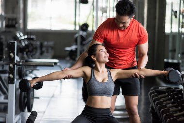 Spor salonunda Asyalı genç bayana Dumbbell egzersizi yaptıran kişisel antrenör. Bankta çalışan gülümseyen kız ve mutlu ortağına bak. Vücut geliştirme ve çiftlerin sağlıklı yaşam tarzı.