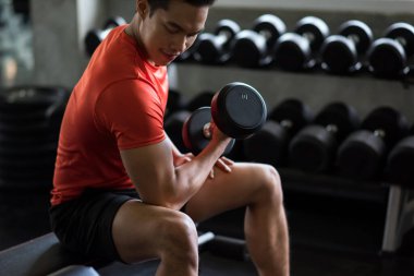 Kas Asyalı gülümseyen adam spor salonunun rafında ağır dambıllarla egzersiz yapıyor. Spor fitness merkezinde kaslı vücut sporcusu. Vücut geliştirme ve sağlıklı yaşam tarzı.