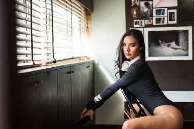 Çekici uzun saçlı, bronz tenli, kulaklıklı Asyalı kadın portresi pencere kenarındaki doğal ışıkla sandalyede oturuyor. Seksi genç kız oturma odasında rahatla..