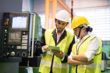 Waist up Asian factory workers or inspectors with safety helmet check corporate app in digital tablet near monitor of CNC lathe milling machine. Manufacturing industry to electronic parts.