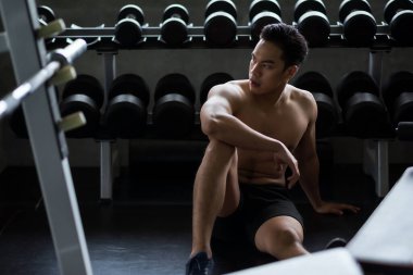 Muscular strong Asian young man with nude torso break and rest after dumbbell bicep exercise near rack in fitness gym. Bodybuilding and healthy sport lifestyle concept.