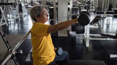 Old senior Asian fit man lifting and stretching dumbbell by right hand in fitness gym with sport equipment bacground. Biceps exercise, bodybuiliding and heatlhy lifestyle for elderly pensioner.