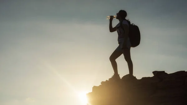 Silhouette hiking woman drink water and stand on top of cliff hill during sunset  after climbing to mountain summit peak. Extreme outdoor adventure sport and bodybuilding healthy lifestyle.