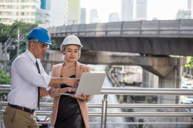 Mühendis ekip çalışması modern şehirdeki inşaat sahasında proje planını tartıştı. Genç Amerikalı bayan mühendis ve kıdemli patron dizüstü bilgisayardaki planlara bakıyorlar..