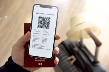 Coronavirüs çağında tatil. Bir adamın elinde Covid-19 'a karşı dijital sertifikalı bir cep telefonu ve seyahat çantasının yanında bir pasaport var..