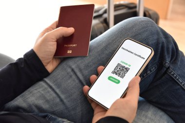 Covid-19 'a karşı dijital aşı sertifikası. Adamın biri seyahat çantasının yanında bir pasaport ve cep telefonu tutuyor. Coronavirus salgını sırasında seyahat kavramı.
