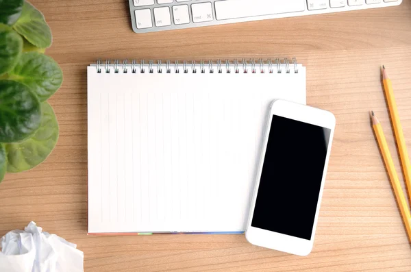 Tampilan top ruang kerja dengan smartphone melalui notepad kosong, persediaan, bunga dan komputer pada latar belakang meja kayu — Stok Foto