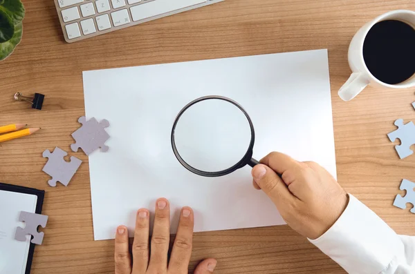 Primo piano della lente d'ingrandimento mano uomo d'affari su carta bianca copia spazio e pezzi di puzzle sparsi sul tavolo — Foto Stock