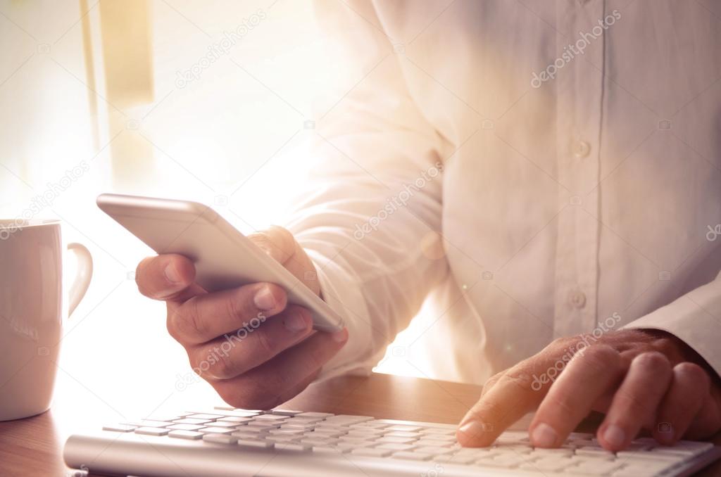 Man's hands using smart phone and computer at office desk