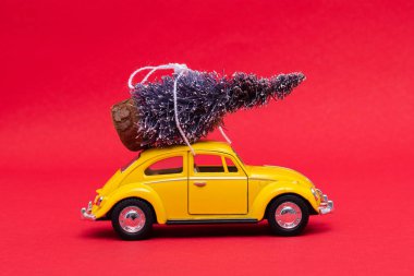 Sarı oyuncak araba kırmızı arka planda, çatıda şenlikli Noel ağacı taşıyor.
